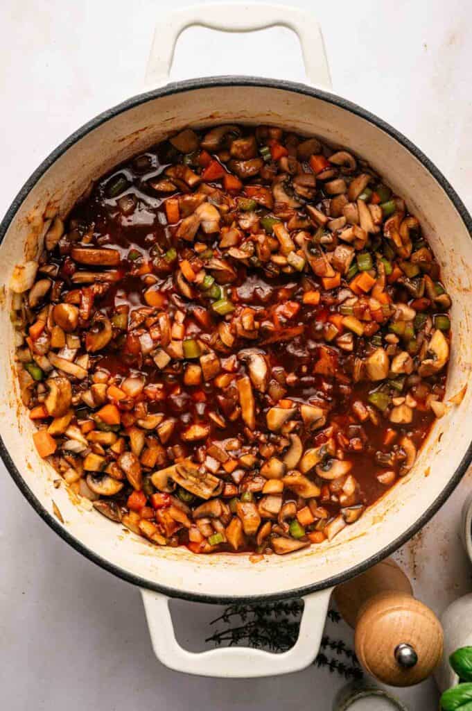A white pot filled with chopped vegetables and mushrooms simmering in a rich, tomato-based sauce, creating a delicious Mushroom Bolognese on a white surface.
