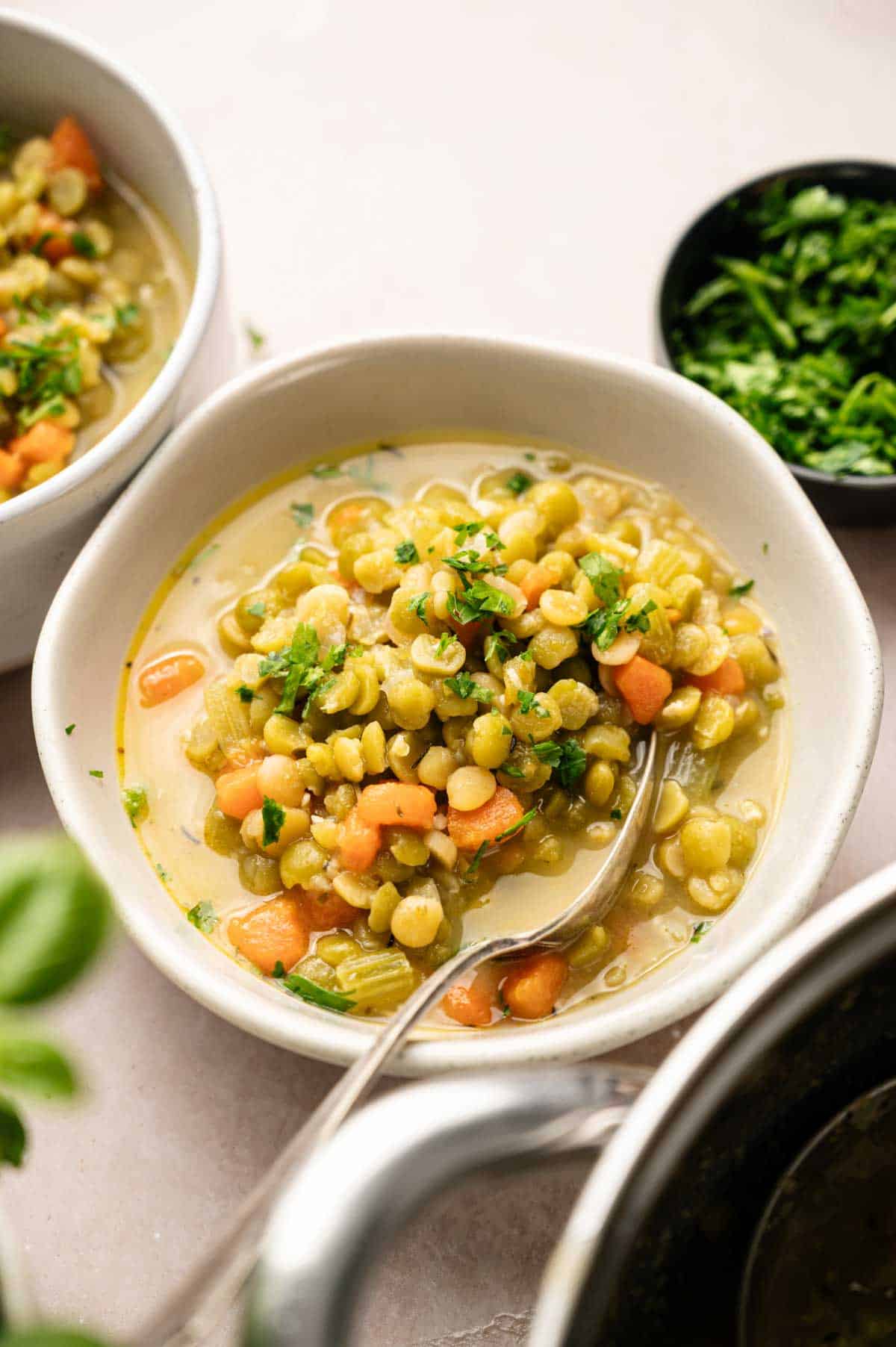 A bowl of Split Pea Soup with diced carrots and celery, garnished with chopped parsley, with a spoon in the bowl and another bowl and fresh herbs visible in the background.