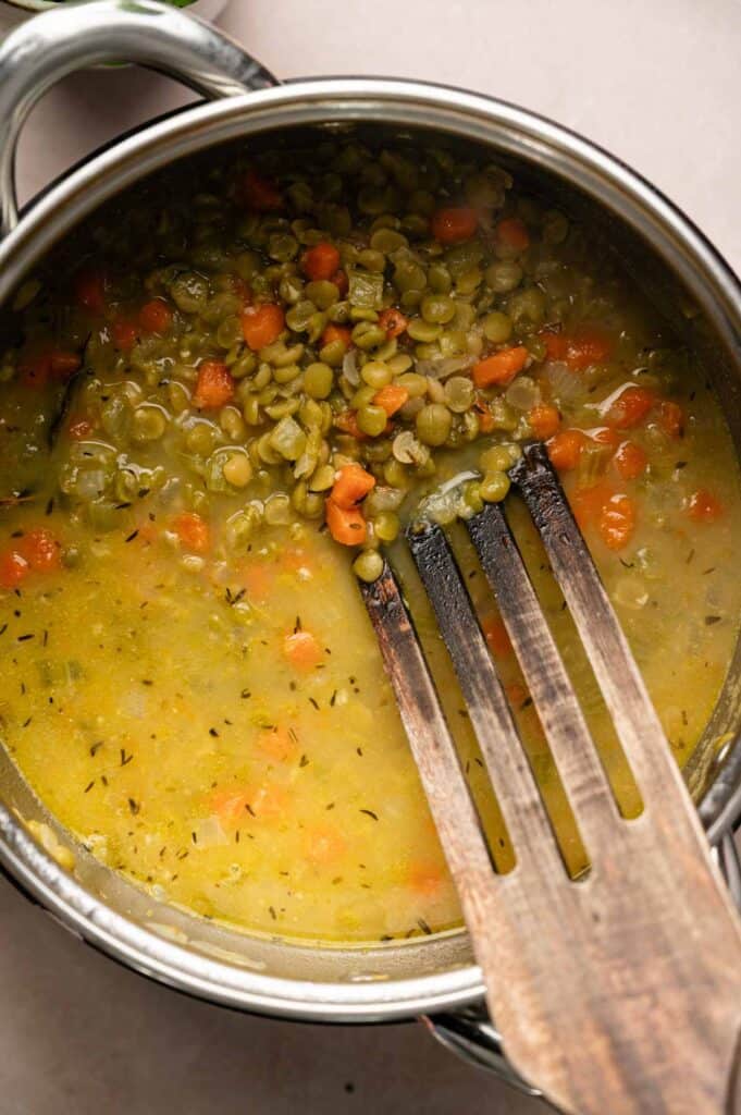 A pot of Split Pea Soup with chopped carrots and peas, partially stirred with a wooden spatula.