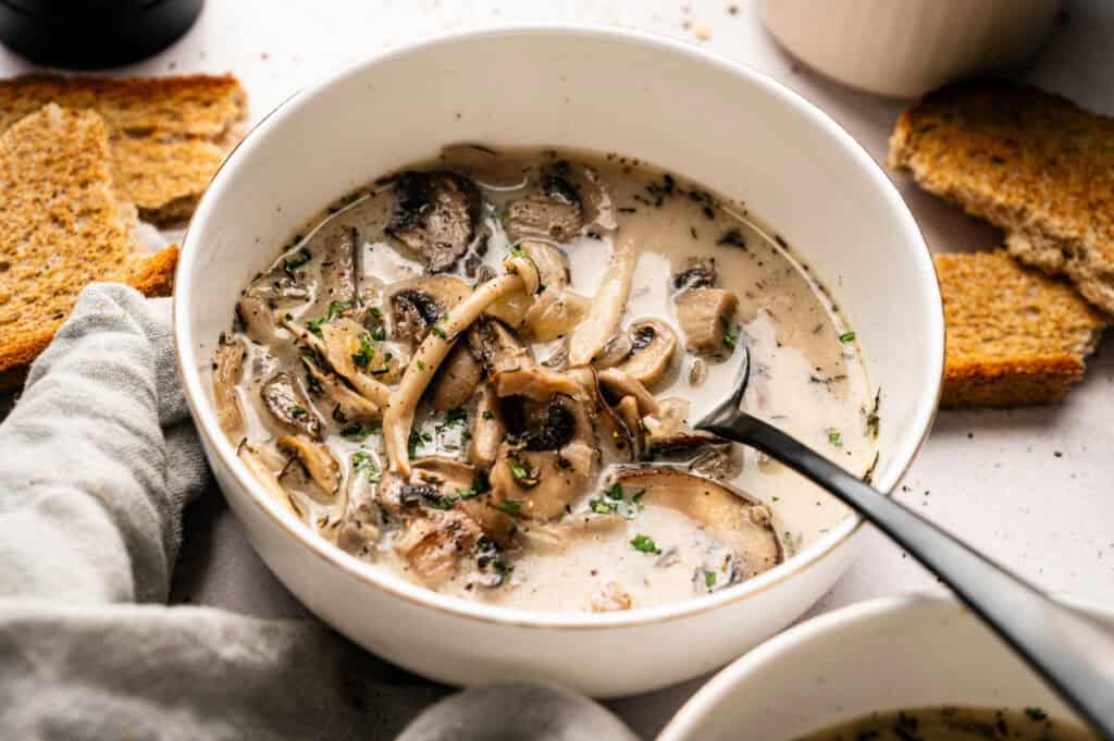 A bowl of creamy Vegan Mushroom Soup with sliced mushrooms and herbs, served with toasted bread on the side.