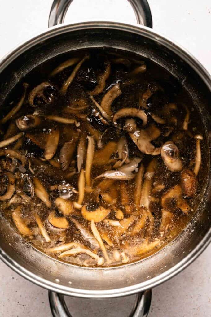 A stainless steel pot filled with Vegan Mushroom Soup, featuring a clear broth and sliced mushrooms, seen from above.