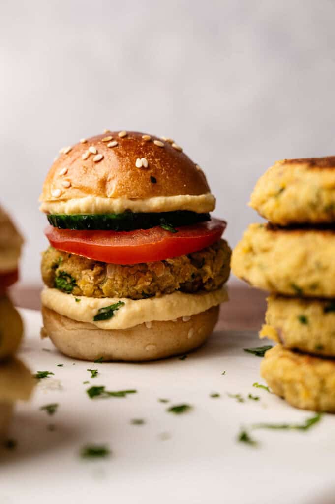 A delicious plate featuring Vegan Sliders: a veggie burger with tomato, cucumber, and sauce on a sesame bun sits on a white surface next to a stack of patties.