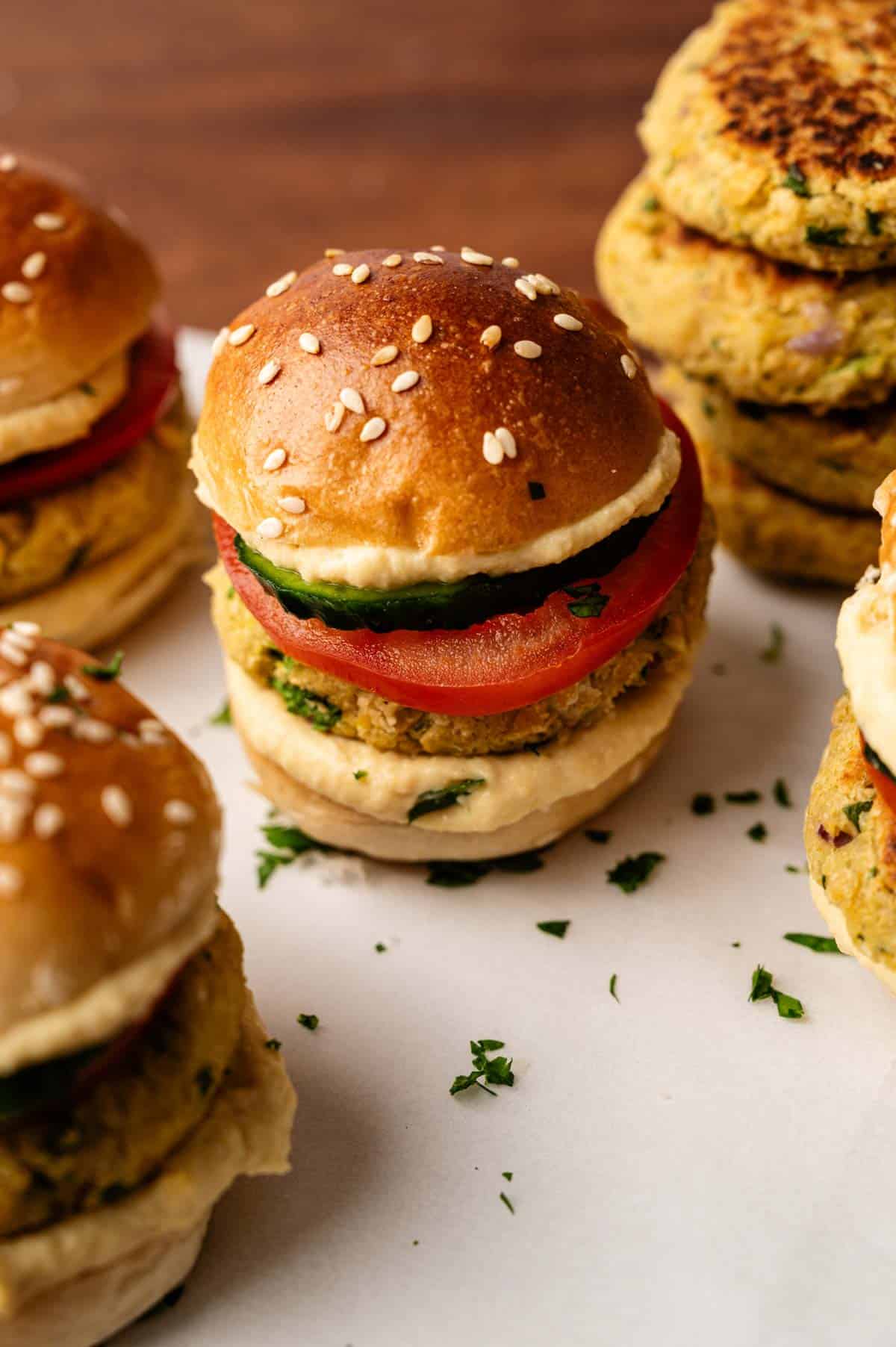 A close-up of vegan sliders with sesame buns, tomato slices, spinach, and a patty, arranged on a white surface with scattered parsley.