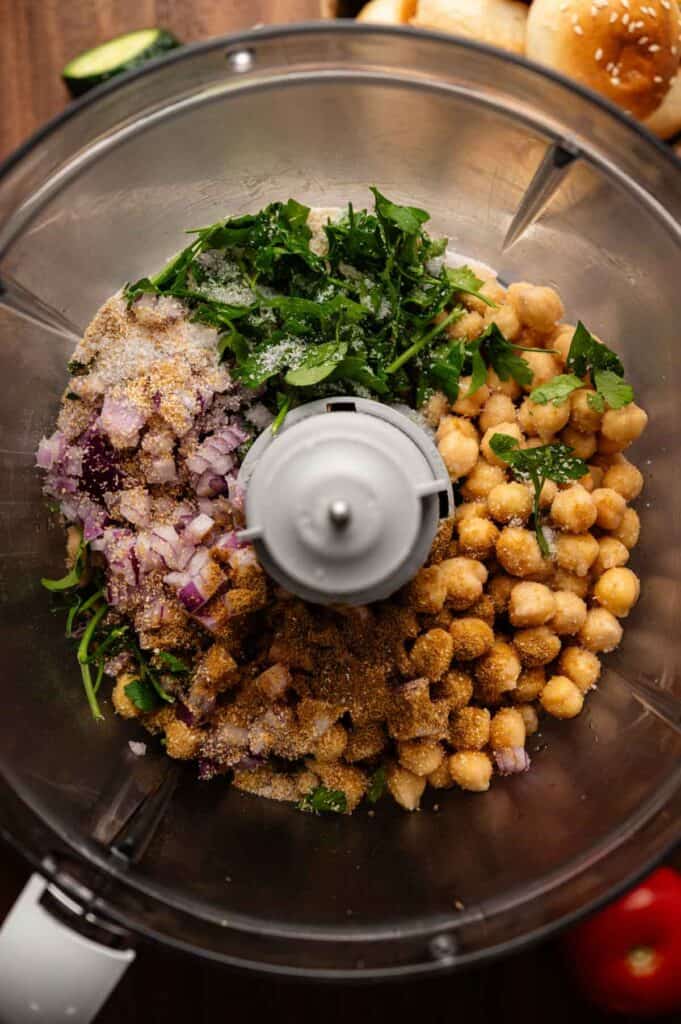 Chickpeas, chopped red onion, parsley, spices, and breadcrumbs in a food processor bowl, ready to be blended for delicious Vegan Sliders.