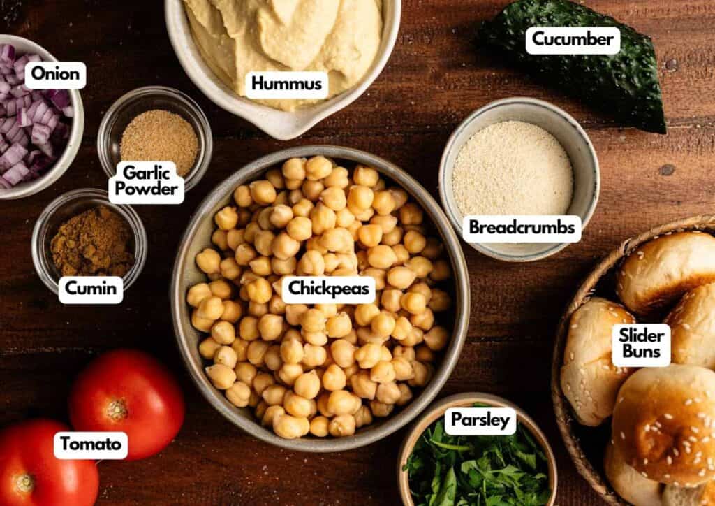 A flat lay of labeled ingredients for making Vegan Sliders, featuring chickpeas, hummus, breadcrumbs, cucumber, slider buns, parsley, tomato, cumin, onion, and garlic powder.
