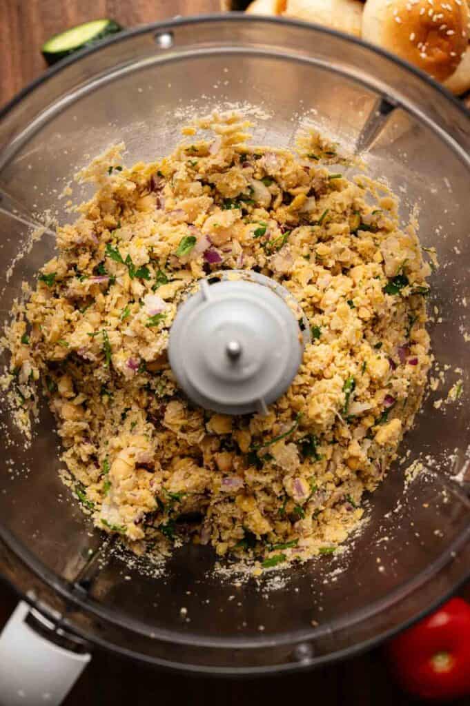 A food processor containing a chunky mixture of chickpeas, chopped red onions, herbs, and seasonings, ready for making falafel or shaping into tasty vegan sliders.