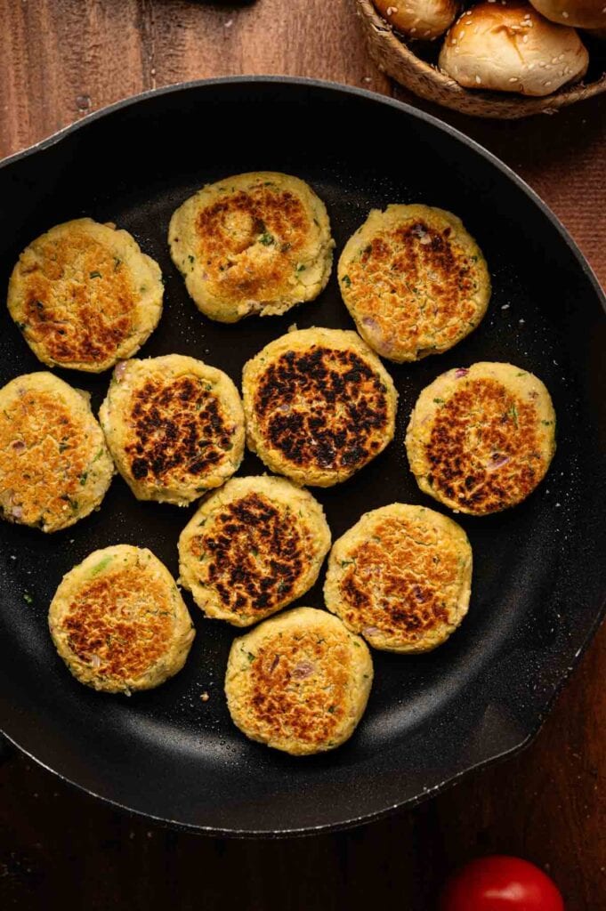 A black skillet with eleven golden-brown vegan sliders cooking on a wooden surface, with mushrooms visible in a basket nearby.
