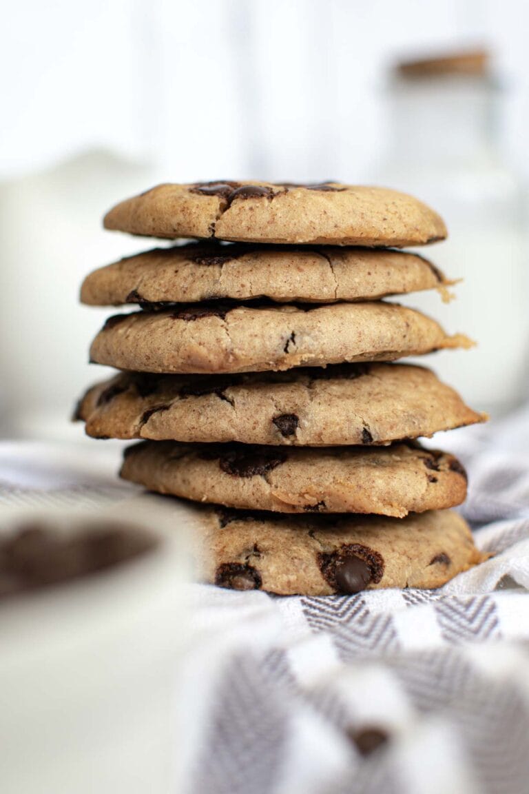 A stack of five White Bean Chocolate Chip Cookies rests on a striped cloth, with a blurred glass of milk and a jar in the background.