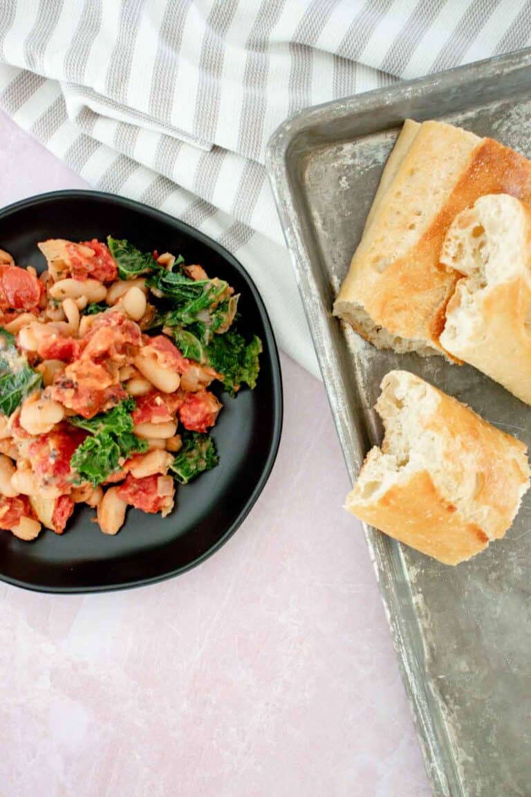 A black plate with a Tuscan White Bean Skillet featuring kale and tomatoes sits next to torn pieces of baguette on a metal tray, with a striped cloth in the background.