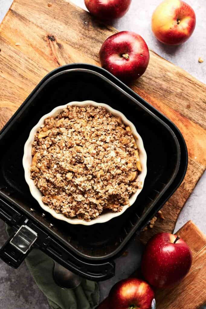 A round baking dish with Air Fryer Vegan Apple Crisp sits in an air fryer basket on a wooden surface, surrounded by whole red apples.