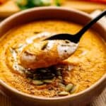A bowl of creamy Vegan Carrot Soup garnished with pumpkin seeds, black pepper, and a swirl of cream, with a spoon lifting a portion.