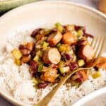 A bowl of white rice topped with sliced sausage, kidney beans, and chopped celery brings classic Vegan Gumbo flavors to your table, served with a gold fork on the side.