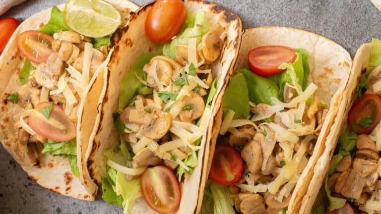 Three soft tacos filled with lettuce, sliced mushrooms, shredded cheese, and halved cherry tomatoes, garnished with a lime wedge on a gray surface.