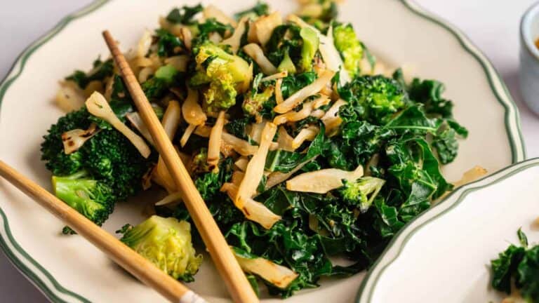 A plate of stir-fried kale, broccoli, and sliced onions with a pair of wooden chopsticks resting on the side.