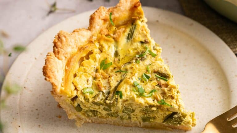 A slice of vegetable quiche with a golden crust, topped with chopped herbs, served on a beige plate with a fork.