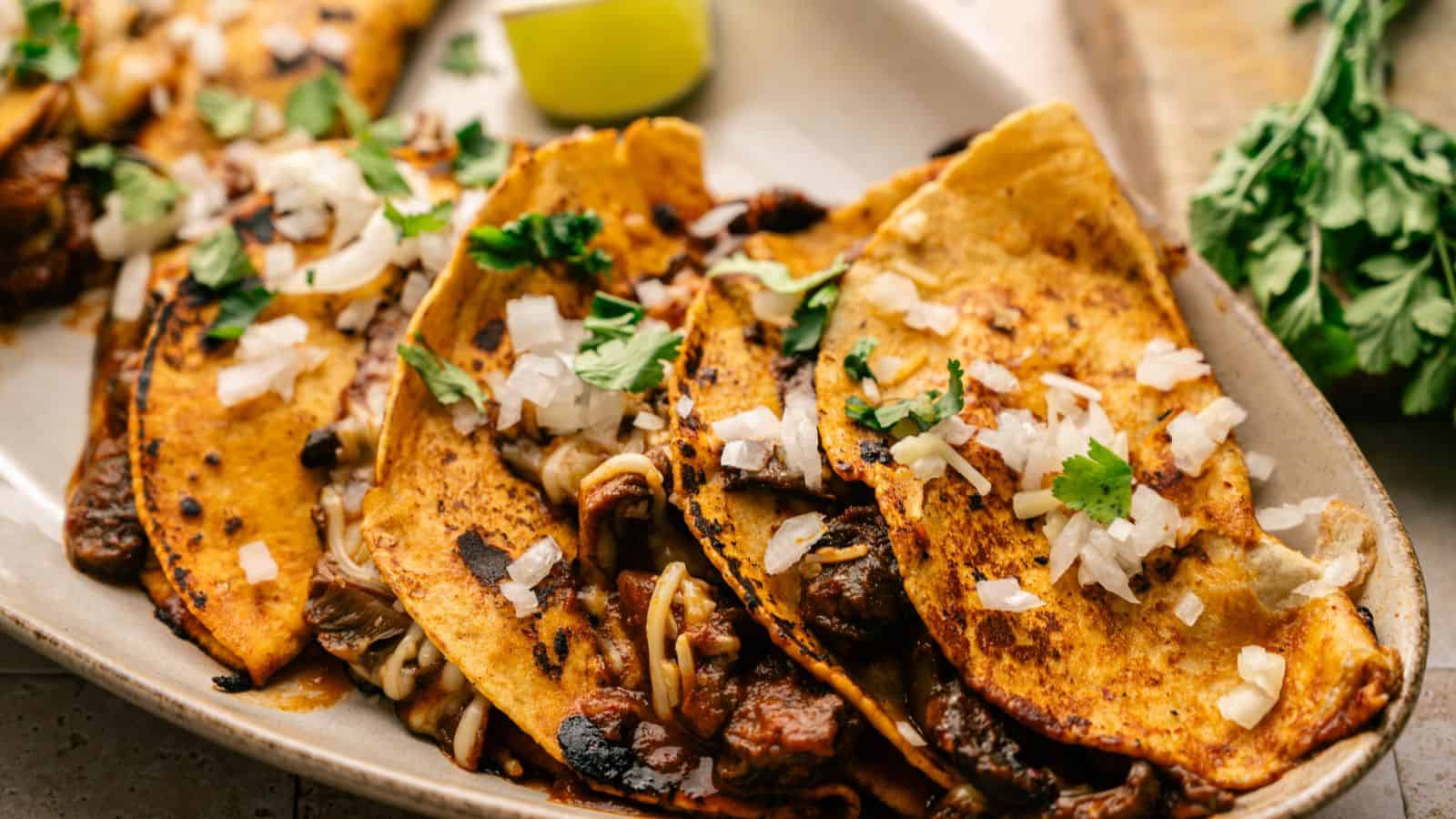 A close-up of three birria tacos on a plate, topped with chopped onions and cilantro, with a lime wedge and fresh herbs in the background.