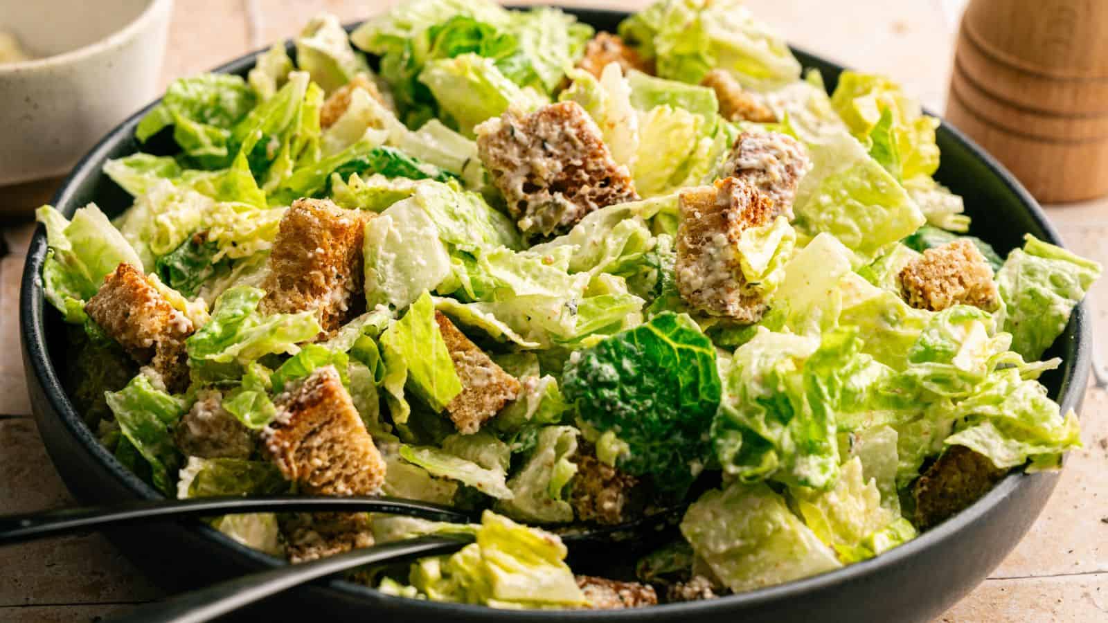A bowl of Caesar salad with romaine lettuce, croutons, and creamy dressing, with serving tongs resting in the salad.