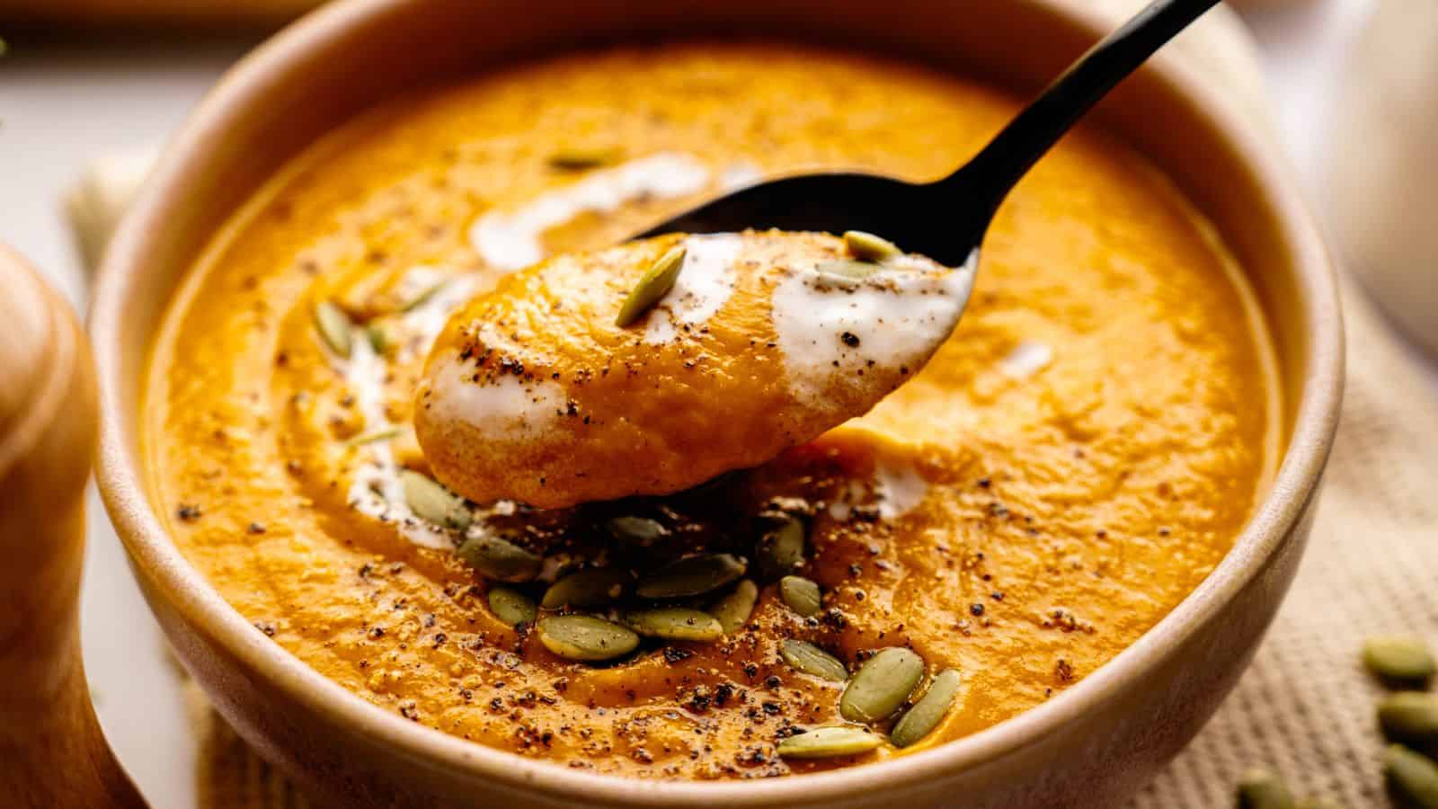 A bowl of creamy orange soup topped with pumpkin seeds, ground pepper, and a swirl of cream, with a spoonful lifted above the bowl.
