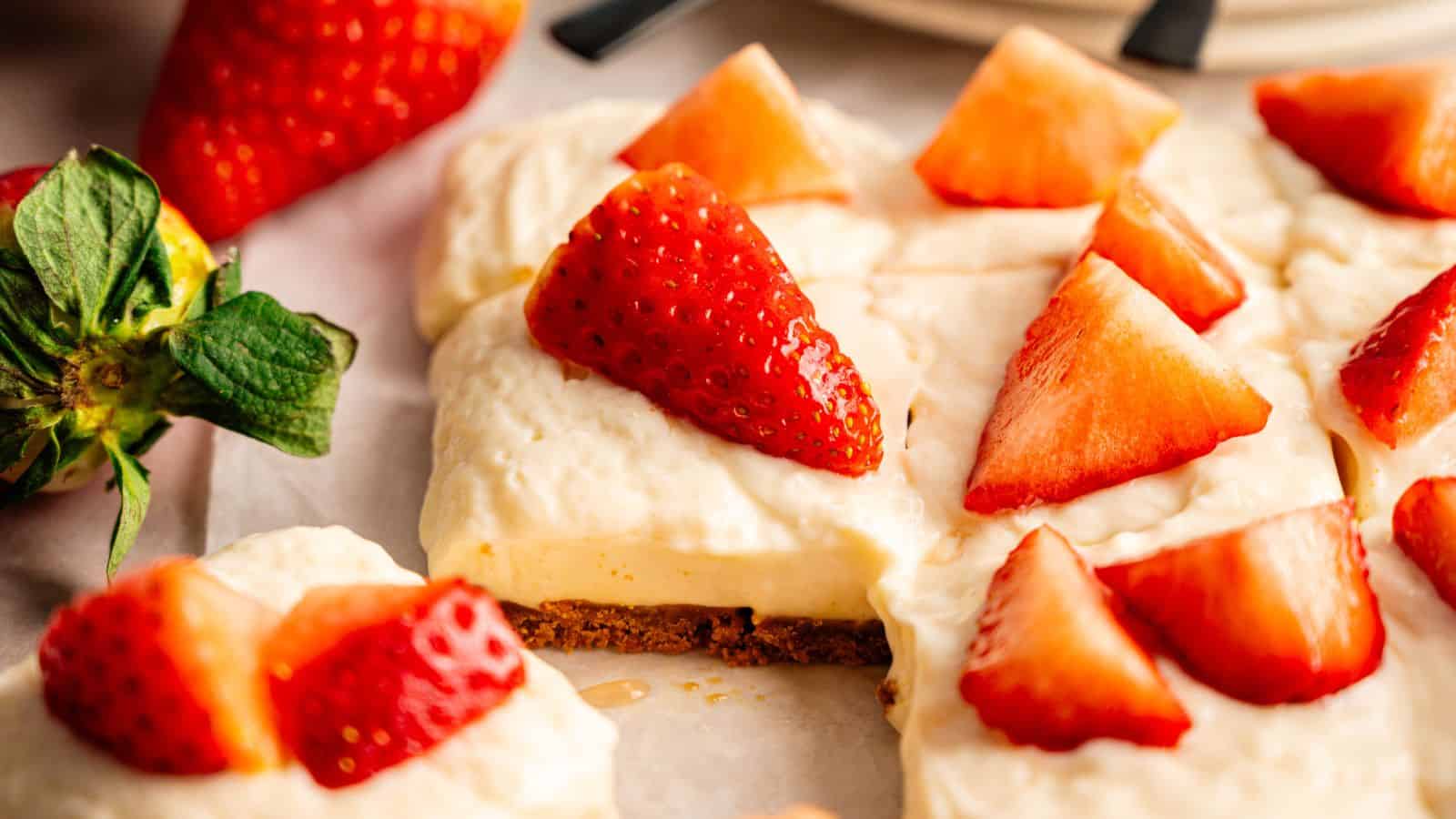 A close-up of a creamy dessert topped with fresh strawberry slices on a biscuit base, with whole and sliced strawberries scattered around.