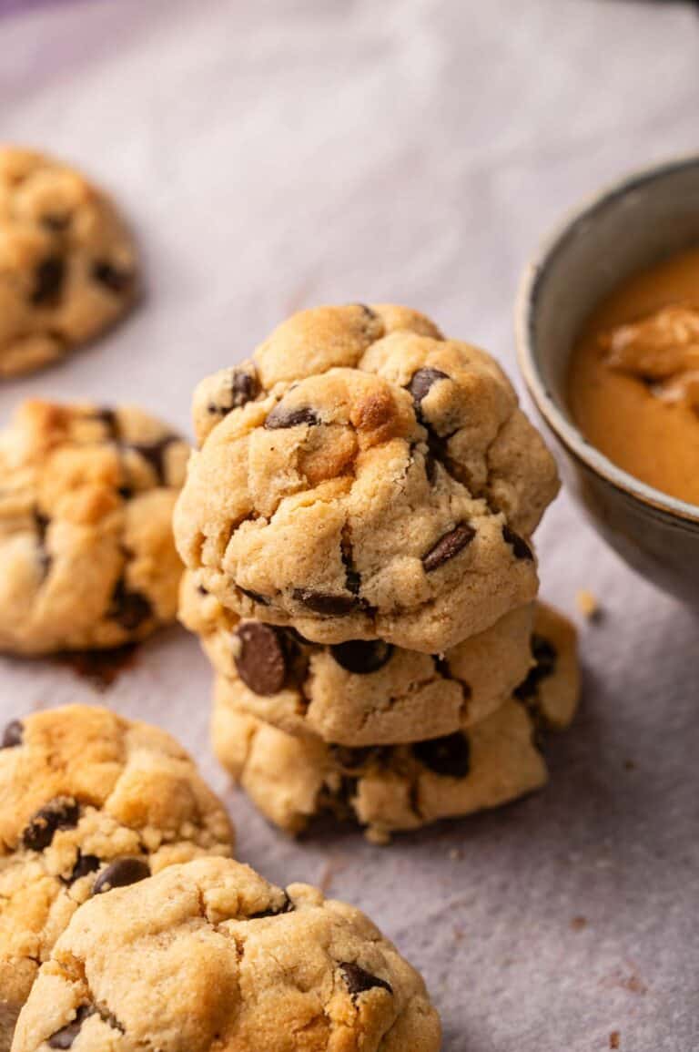 A stack of chocolate chip cookies on parchment paper next to a bowl of peanut butter, with more cookies scattered around.