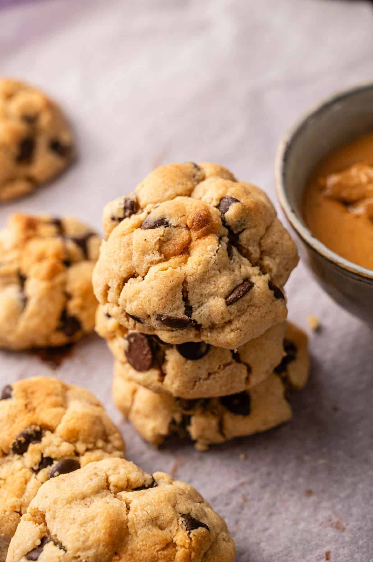 A stack of chocolate chip cookies on parchment paper next to a bowl of peanut butter, with more cookies scattered around.