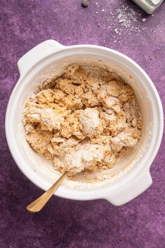 A white mixing bowl with partially mixed Peanut Butter Chocolate Chip Cookies dough and a gold spoon sits on a purple countertop.