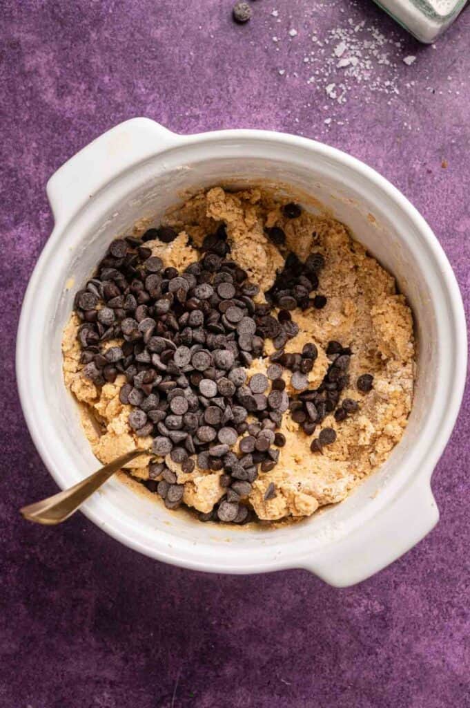 A white mixing bowl with peanut butter chocolate chip cookie dough and chocolate chips, with a spoon in the dough, on a purple countertop.