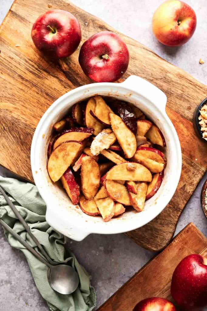 Sliced apples mixed with spices in a white baking dish, surrounded by whole apples, a green cloth, and two spoons on a wooden cutting board—perfect for preparing an Air Fryer Vegan Apple Crisp.