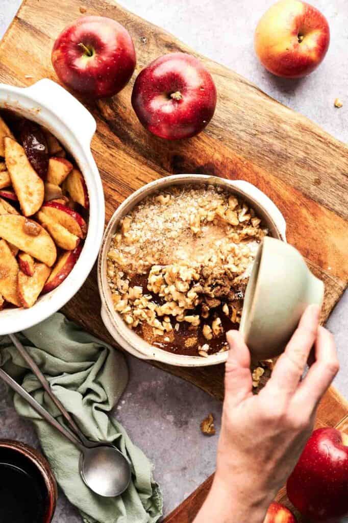 A hand lifts the lid off a round baking dish filled with Air Fryer Vegan Apple Crisp, surrounded by apples and a bowl of sliced apples on a wooden cutting board.