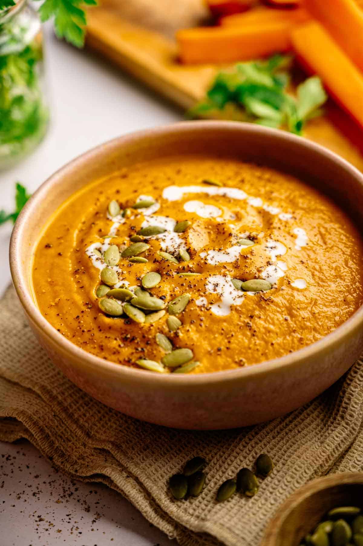 A bowl of creamy orange Vegan Carrot Soup topped with pumpkin seeds, a swirl of cream, and ground pepper, with carrot sticks and herbs in the background.