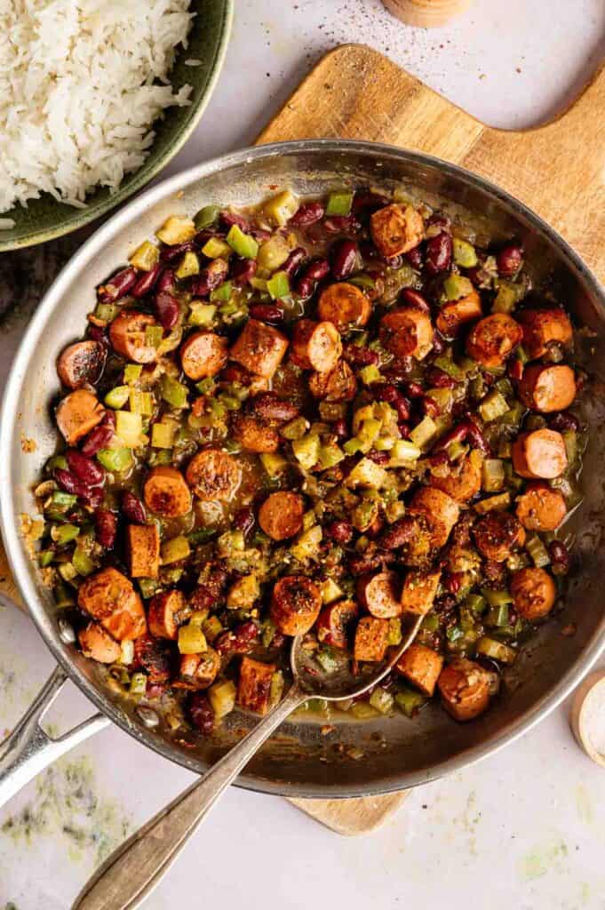 A skillet filled with sliced sausage, kidney beans, chopped green vegetables, and spices creates a hearty vegan gumbo; a spoon rests inside beside a bowl of white rice.