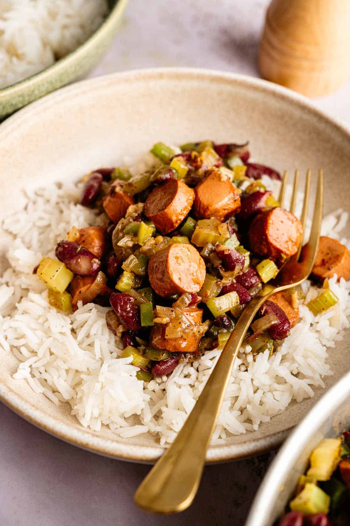 A plate of white rice topped with a hearty Vegan Gumbo—featuring sliced sausage, kidney beans, and chopped vegetables—with a gold fork placed on the side.