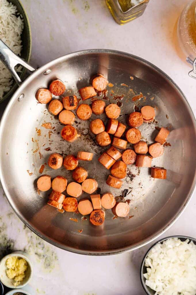 Sliced sausages being browned in a stainless steel skillet, surrounded by bowls of chopped onions, garlic, cooked rice, and other ingredients on a kitchen counter—all the makings for a hearty Vegan Gumbo.