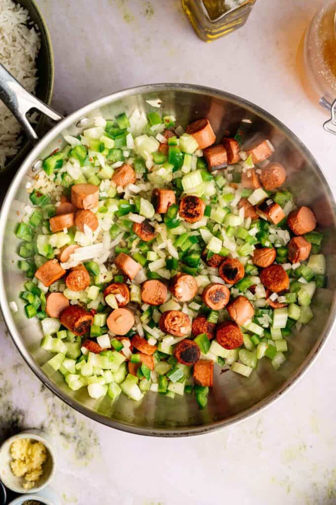 A pan with sliced sausage, chopped celery, green bell pepper, and onions being sautéed on a light kitchen countertop—a perfect start for a hearty Vegan Gumbo.