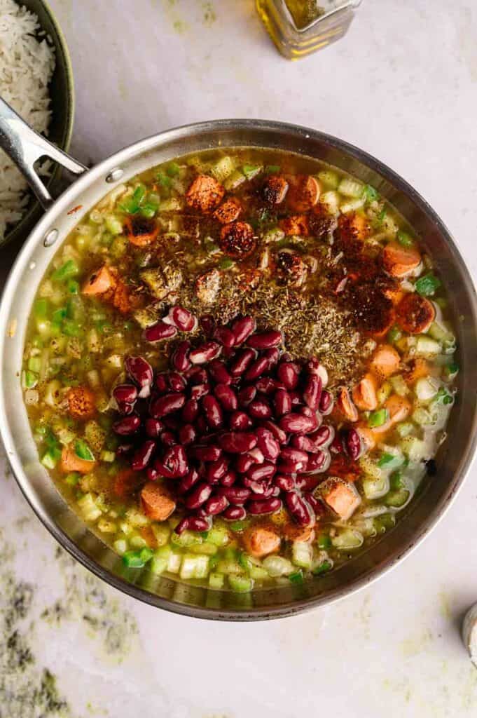 A skillet filled with chopped vegetables, spices, and kidney beans simmering in broth creates a hearty Vegan Gumbo, served next to a bowl of white rice.