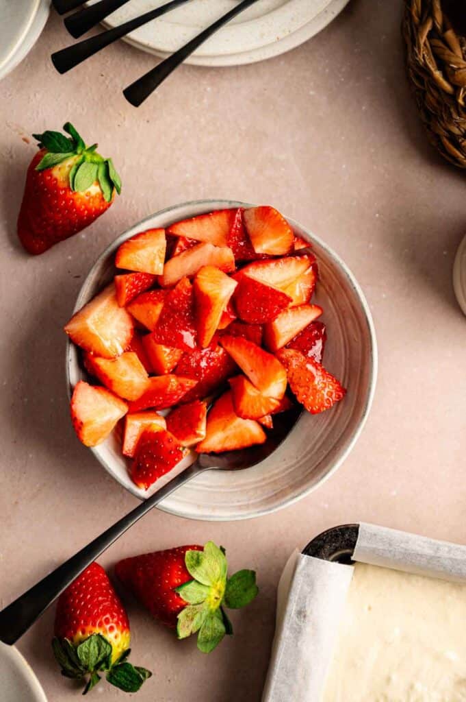 A bowl of sliced strawberries with a spoon inside, surrounded by whole strawberries and plates on a beige surface—perfect for topping your favorite vegan strawberry cheesecake.