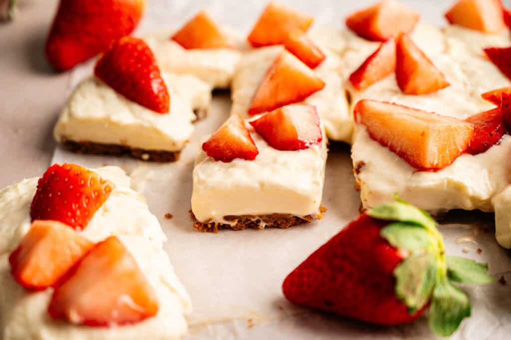 Squares of Vegan Strawberry Cheesecake bars topped with fresh strawberry slices are arranged on parchment paper, with whole and cut strawberries scattered around.