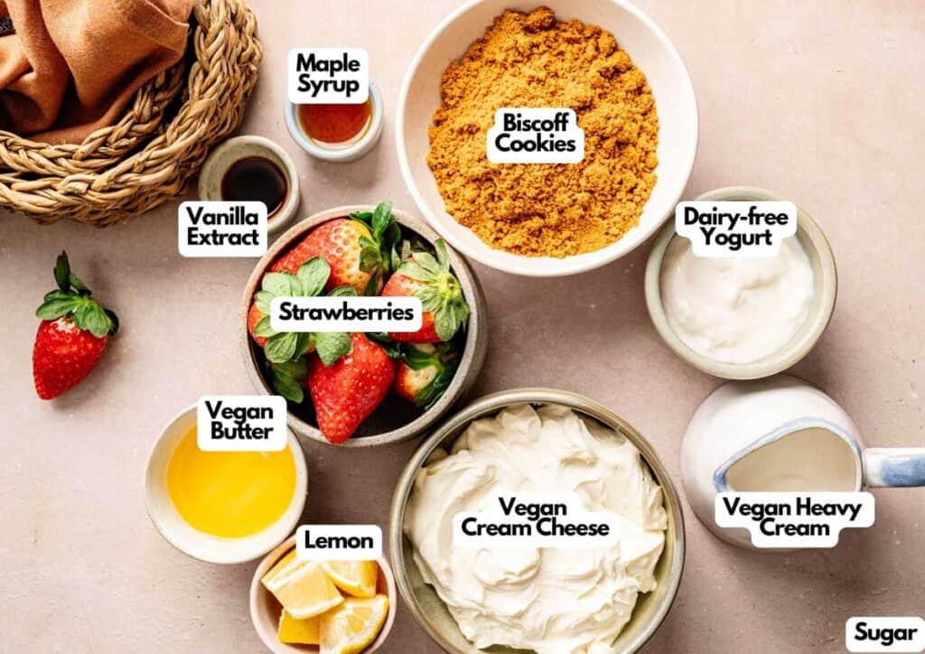 Various labeled ingredients for a Vegan Strawberry Cheesecake, including strawberries, vegan cream cheese, vegan butter, lemon, vanilla extract, maple syrup, biscoff cookies, sugar, dairy-free yogurt, and vegan heavy cream.