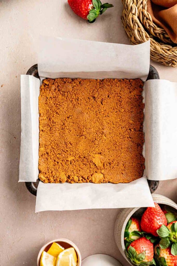A square baking pan lined with parchment paper holds a pressed crumb crust, ready to become a vegan strawberry cheesecake. Surrounding the pan are fresh strawberries and lemon wedges in small bowls.