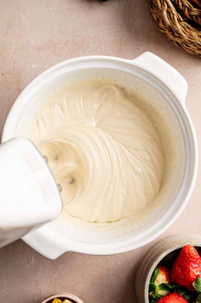 An electric hand mixer blends creamy batter for a Vegan Strawberry Cheesecake in a white bowl on a countertop, with fresh strawberries visible nearby.