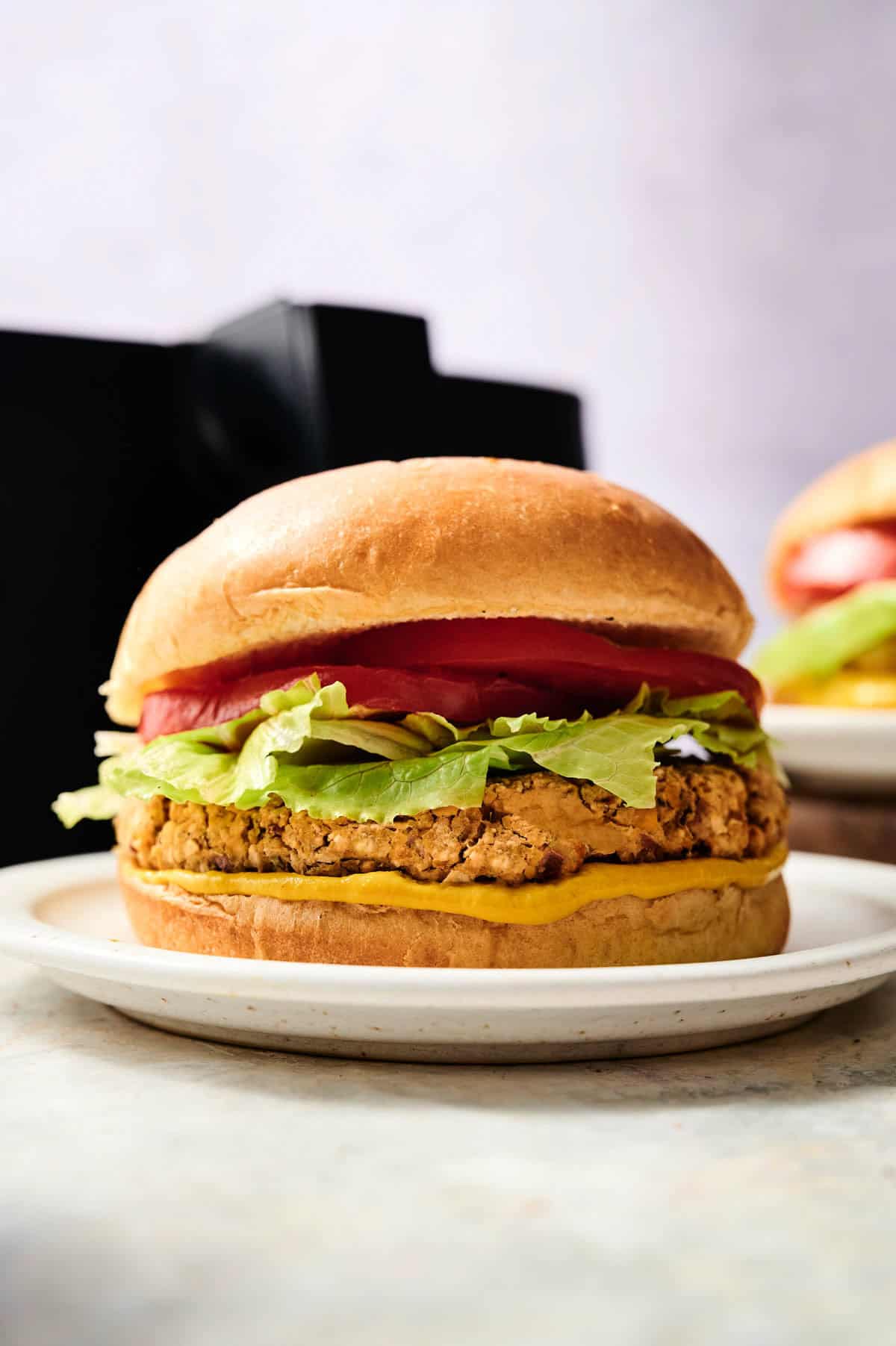 An Air Fryer Chickpea Burger with lettuce, tomato, and mustard on a sandwich bun, served on a white plate.