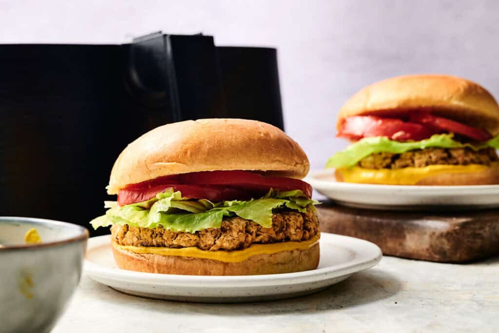 An Air Fryer Chickpea Burger with lettuce, tomato, and mustard on a bun sits on a white plate, with another similar burger in the background.