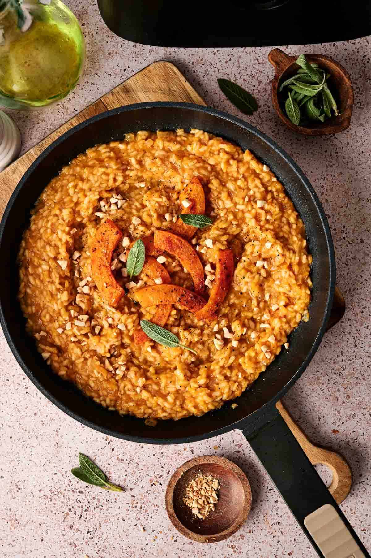 A pan of Air Fryer Pumpkin Risotto garnished with roasted pumpkin slices, chopped nuts, and fresh sage leaves on a light countertop.