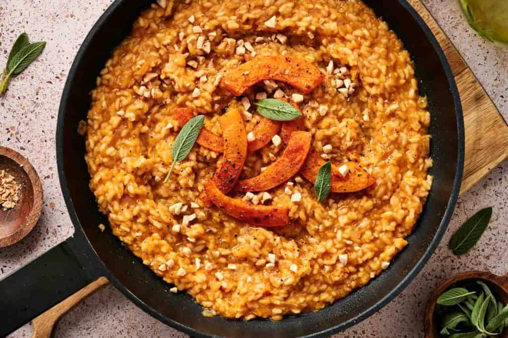 A pan of creamy Air Fryer Pumpkin Risotto garnished with roasted pumpkin slices, chopped nuts, and fresh sage leaves sits on a countertop.