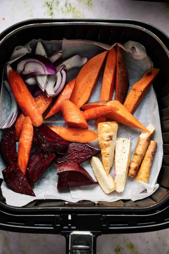Air Fryer Roasted Root Vegetables like carrots, sweet potatoes, parsnips, beets, and red onions are arranged on parchment paper in the basket, ready for delicious roasting.