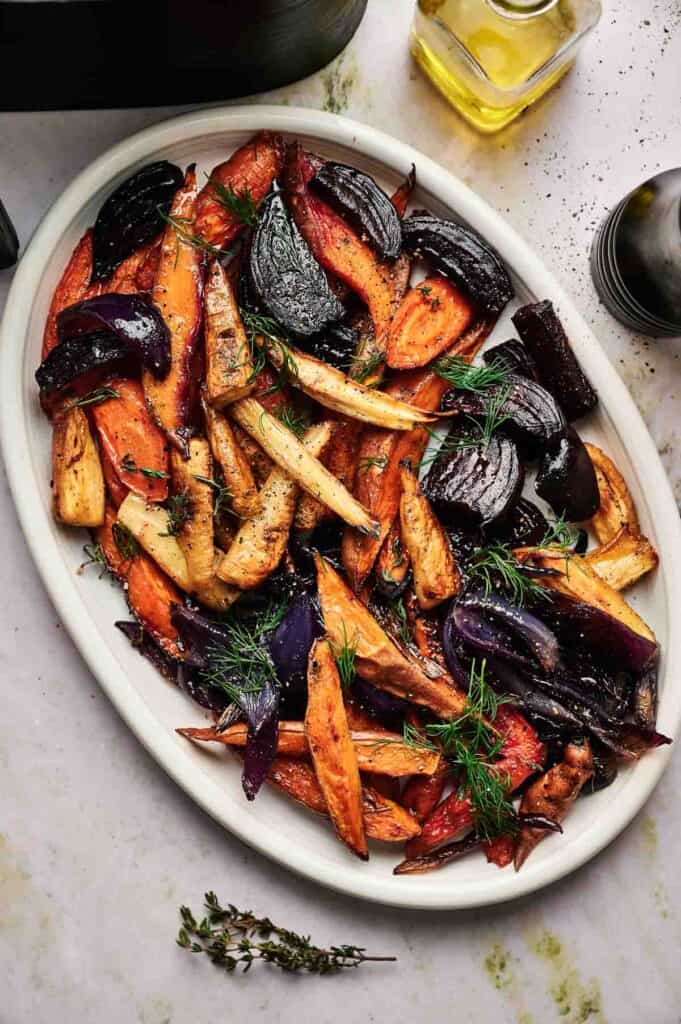 A white oval platter with Air Fryer Roasted Root Vegetables, including carrots, parsnips, and purple onions, garnished with fresh herbs on a marble surface.