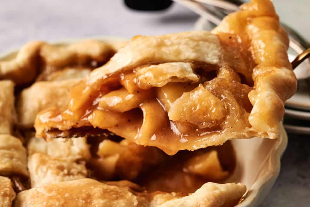A close-up of a slice of Air Fryer Vegan Apple Pie being lifted from a pie dish, showing its golden crust and cinnamon-coated apple filling.