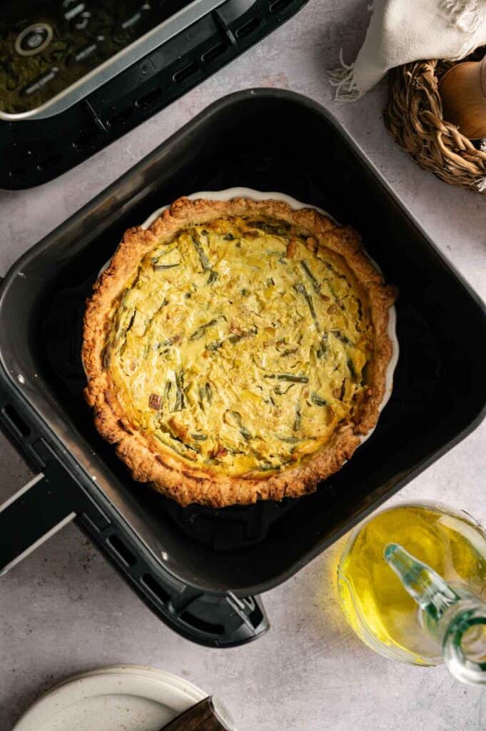 An Air Fryer Vegan Asparagus Quiche with a golden crust sits in an air fryer basket. A bottle of olive oil, eggs in a basket, and a cloth napkin are nearby on the countertop.