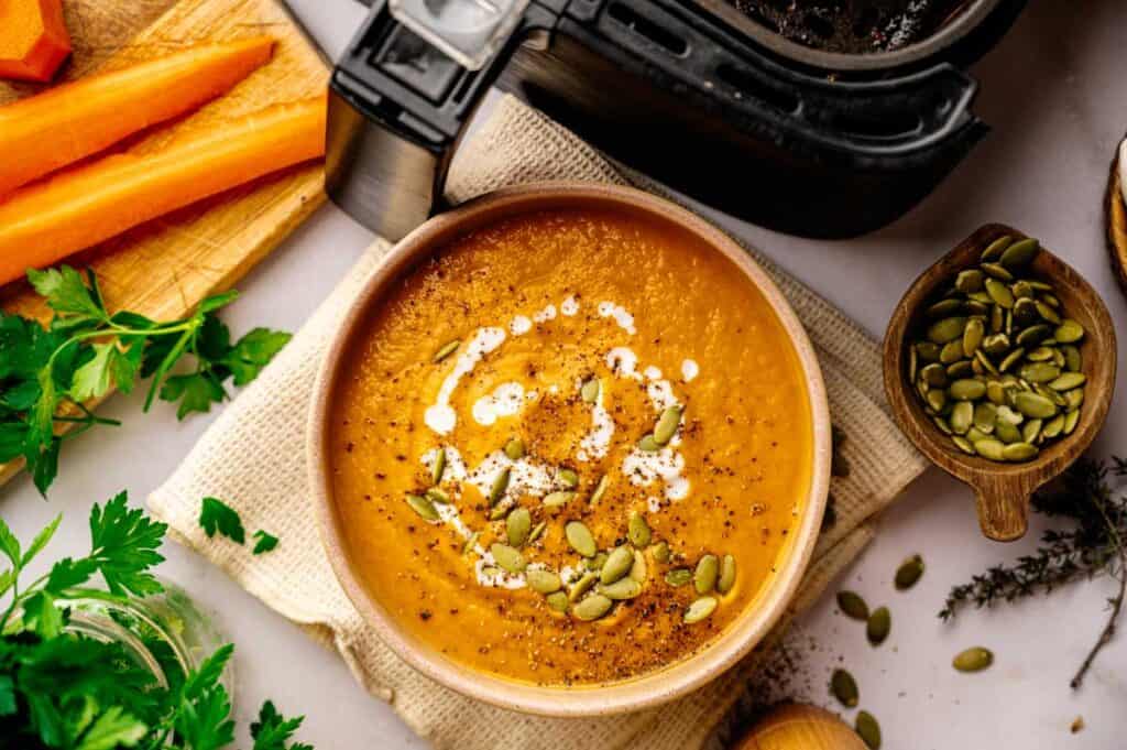 A bowl of creamy orange Air Fryer Vegan Carrot Soup topped with a swirl of cream and pumpkin seeds, placed next to an air fryer, fresh herbs, carrot sticks, and a wooden spoon.