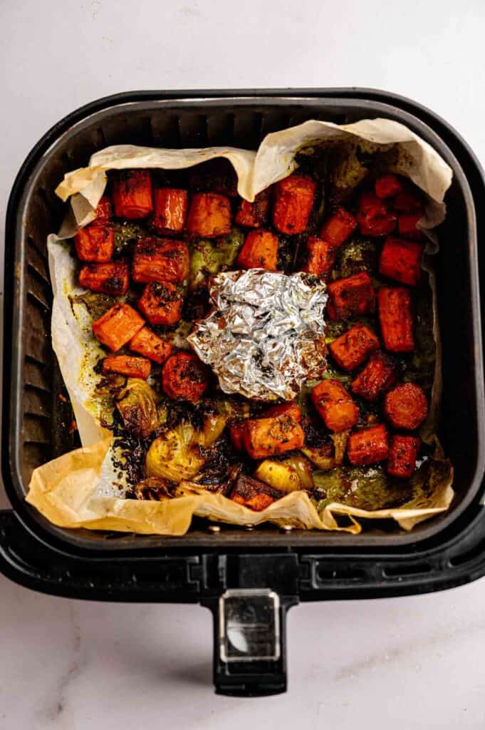 Chopped seasoned carrots and onions are roasted in an air fryer lined with parchment paper for a delicious base to Air Fryer Vegan Carrot Soup; a small piece is wrapped in aluminum foil at the center.